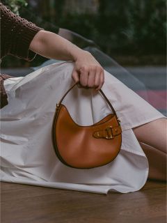 Eine Person in einem weißen Rock hält eine braune Lederhandtasche mit einer Schnalle und sitzt in einem Haus in der Nähe eines Fensters.