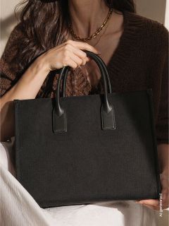 Eine Frau in einem braunen Pullover hält eine große schwarze Handtasche mit zwei Henkeln und hat eine weiße Decke auf dem Schoß.