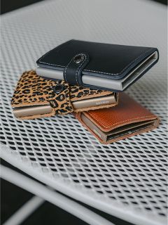 Drei Geldbörsen - schwarz, mit Leopardenmuster und braun - liegen auf einem weißen Metallgittertisch.