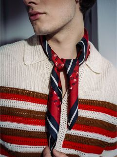 Eine Person, die ein cremefarbenes Strickhemd mit roten und braunen Streifen trägt und einen rot gemusterten Schal um den Hals gebunden hat.