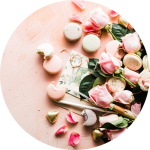 Rosa Rosen, grüne Blätter und verschiedene Macarons sind auf einer hellrosa Fläche arrangiert, mit verstreuten Blütenblättern und einem Paar Gläser in der Nähe.