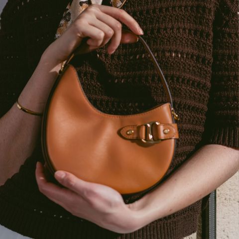Person mit einer kleinen, hellbraunen Lederhandtasche mit Schnallenverschluss, einem braunen Strickoberteil und einem goldenen Armband.