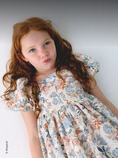 Junges Mädchen mit langen roten Haaren in einem geblümten Kleid steht vor einem weißen Hintergrund und macht ein verspieltes, schmollendes Gesicht.