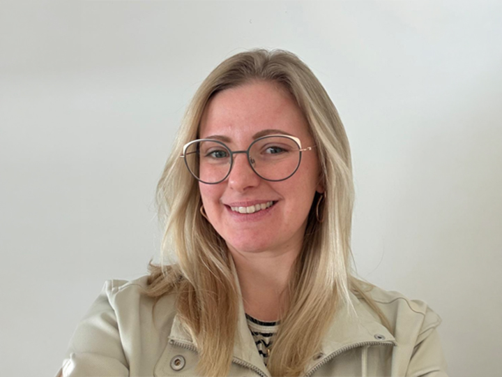 Eine Frau mit langem blondem Haar und Brille lächelt, sie trägt eine helle Jacke und ein gestreiftes Oberteil vor einem schlichten hellen Hintergrund.