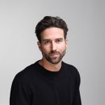 Ein Mann mit kurzen braunen Haaren und Bart, der einen schwarzen Pullover trägt, sitzt vor einem schlichten hellgrauen Hintergrund und schaut mit neutralem Blick in die Kamera.