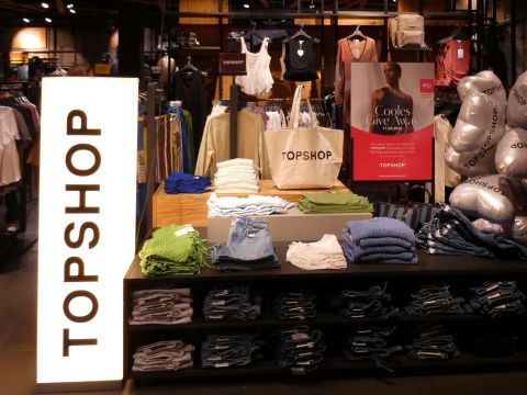 Auslage eines Bekleidungsgeschäfts mit ordentlich gefalteten Oberteilen, Shorts und Taschen in den Regalen, einem hellen "TOPSHOP"-Schild und einem Werbeplakat im Hintergrund.