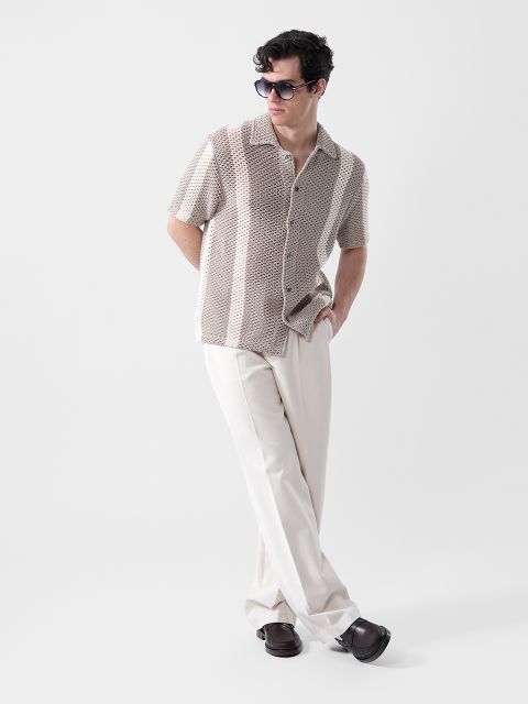 Ein Mann mit Sonnenbrille, einem beigen, gemusterten Kurzarmhemd und einer cremefarbenen Hose steht mit einer Hand in der Tasche vor einem schlichten Hintergrund.