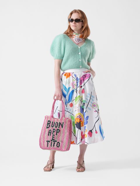 Frau mit Sonnenbrille, die eine mintgrüne Strickjacke, einen geblümten Rock, einen Schal und Sandalen trägt und eine rosa Tragetasche mit der Aufschrift "BUON APPETITO" in der Hand hält.