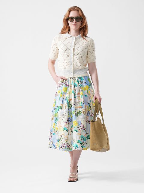 Frau mit Sonnenbrille, cremefarbener, kurzärmeliger Strickjacke, buntem, geblümtem Rock, Sandalen und einer hellbraunen Tragetasche vor weißem Hintergrund.