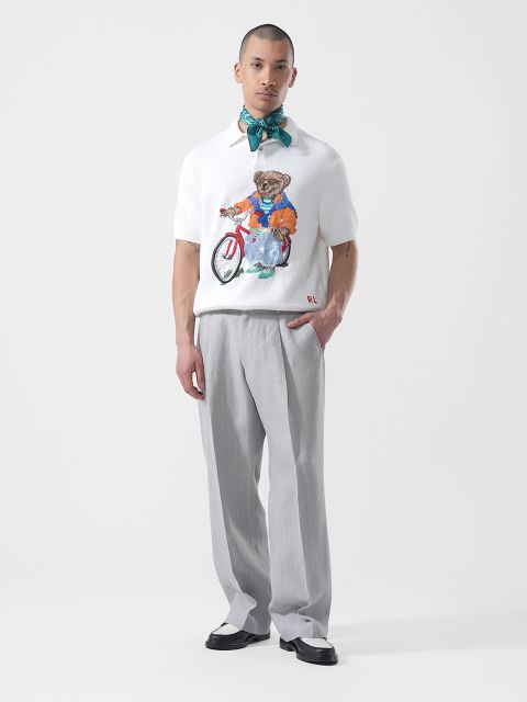 Ein Mann steht in einer hellgrauen Hose, einem weißen Hemd mit einer Bärengrafik, zweifarbigen Schuhen und einem um den Hals gebundenen blaugrünen Schal vor einem weißen Hintergrund.