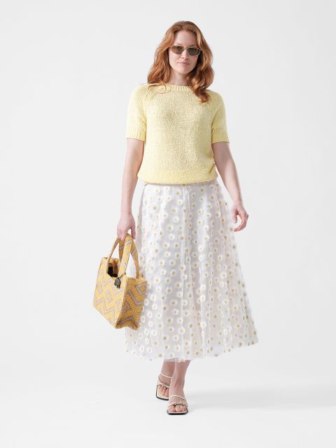 Frau mit Sonnenbrille, die ein gelbes Oberteil, einen weißen Rock mit Gänseblümchenmuster und Sandalen trägt und eine gelb gemusterte Tragetasche vor einem weißen Hintergrund hält.