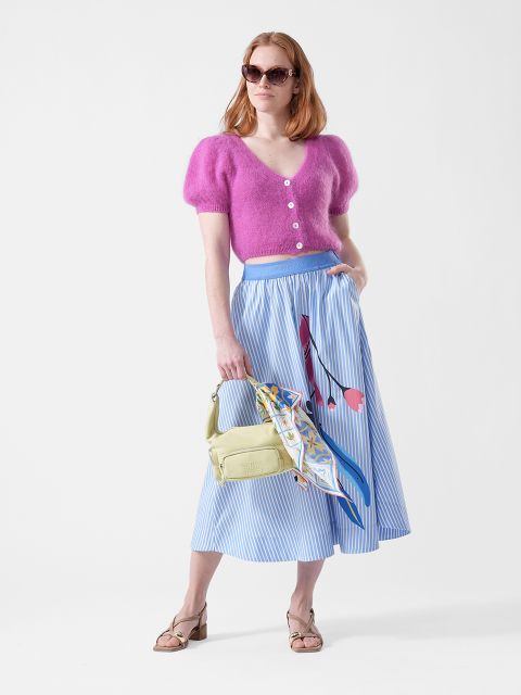 Frau in einem rosafarbenen, wuscheligen Oberteil, einem gestreiften blauen Midirock mit Blumenmuster, einer Sonnenbrille, einer gelben Tasche mit Schal und Sandalen.