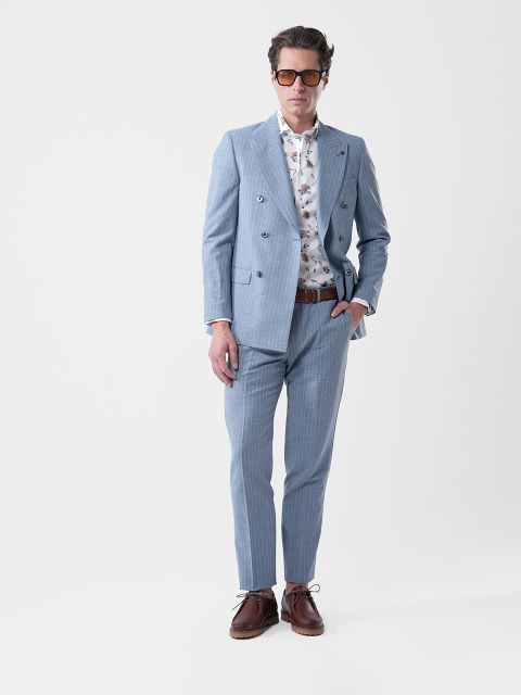 Mann in hellblauem Anzug, gemustertem weißen Hemd, braunen Schuhen und Sonnenbrille, der mit einer Hand in der Tasche vor einem schlichten Hintergrund steht.