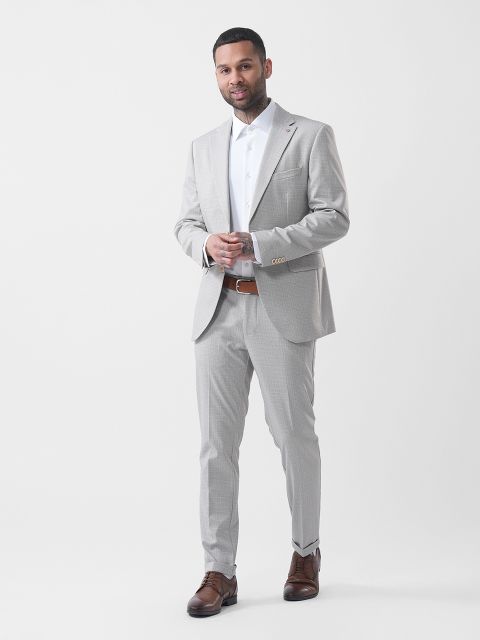 Mann in einem hellgrauen Anzug mit weißem Hemd und braunen Schuhen, der vor einem schlichten weißen Hintergrund posiert.