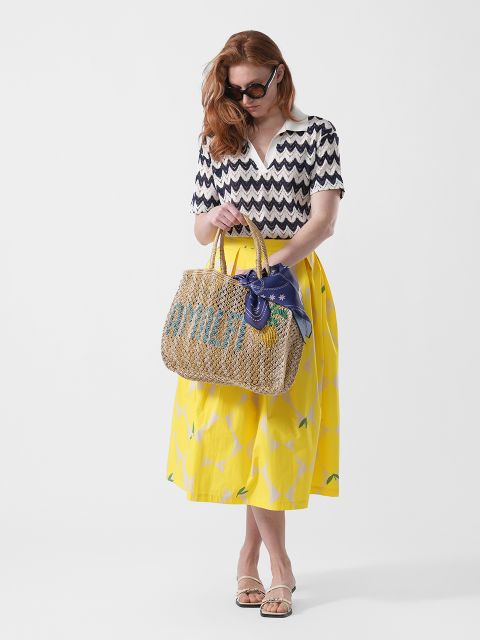 Frau mit Sonnenbrille, die ein zickzackförmiges Oberteil, einen gelben Rock und Sandalen trägt und eine gewebte Tasche mit einem blauen Schal in der Hand hält, steht vor einem weißen Hintergrund.