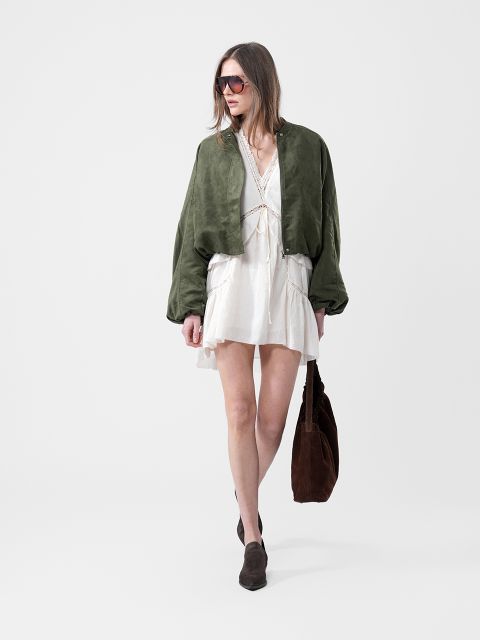 Frau in weißem Kleid, grüner Jacke, Sonnenbrille und braunen Stiefeln, die eine große braune Tasche vor einem schlichten weißen Hintergrund trägt.