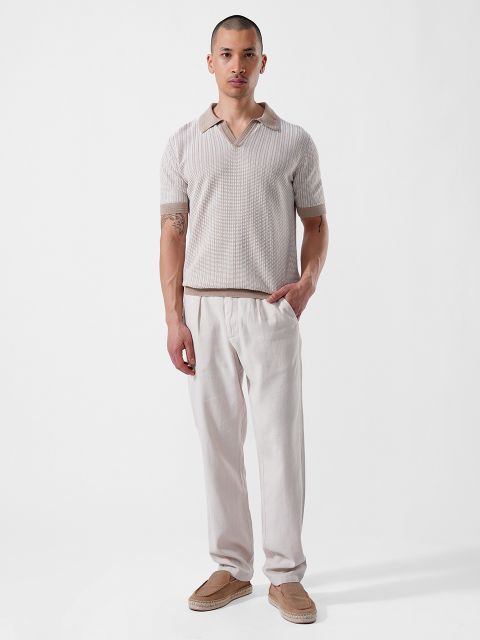 Ein Mann steht vor einem weißen Hintergrund und trägt ein kurzärmeliges, beigefarbenes Hemd mit Kragen, eine helle Hose und hellbraune Slipper.