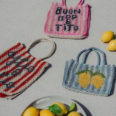 Drei bunte gewebte Taschen, zwei mit italienischem Text und eine mit Zitronen, stehen auf einer hellen Fläche neben einem Teller mit Zitronen und Basilikum.