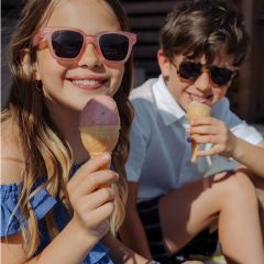 Zwei lächelnde Kinder mit Sonnenbrillen genießen an einem sonnigen Tag Eiswaffeln im Freien.