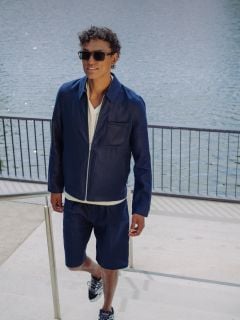 Ein Mann mit Sonnenbrille, marineblauer Jacke und passenden Shorts geht eine Freitreppe an einem Gewässer hinauf, hält sich an einem Geländer fest und lächelt.