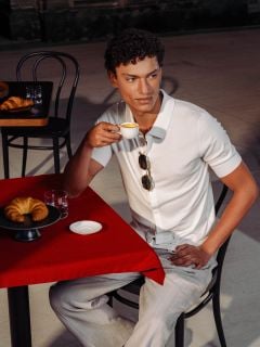 Mann in weißem Hemd sitzt an einem roten Tisch, trinkt Espresso, hat ein Croissant und ein Glas Wasser vor sich in einem Straßencafé.