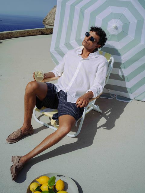 Mann mit Sonnenbrille und weißem Hemd entspannt sich auf einem Liegestuhl mit einem Getränk im Schatten eines gestreiften Sonnenschirms; Zitronen und ein Glas stehen in der Nähe auf dem Boden.