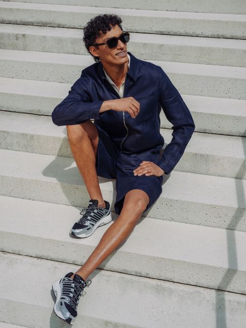 Ein Mann mit Sonnenbrille, marineblauer Jacke und passenden Shorts sitzt auf einer Betontreppe im Freien, lächelt, hat ein Bein angewinkelt und trägt Sportschuhe.