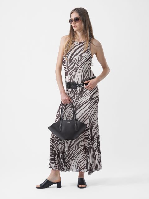 Frau mit Sonnenbrille, die ein schwarz-weiß gemustertes Maxikleid, einen schwarzen Gürtel und schwarze Sandalen trägt und eine schwarze Handtasche vor einem weißen Hintergrund hält.