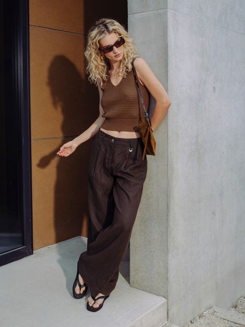 Eine Frau mit lockigem blondem Haar und Sonnenbrille trägt ein braunes ärmelloses Oberteil, weite Hosen und Sandalen und steht vor einem modernen Gebäude.