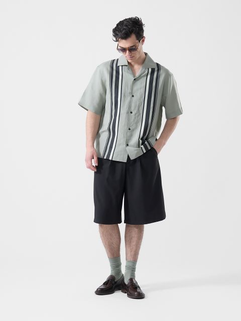 Ein Mann, der eine Sonnenbrille, ein hellgrünes kurzärmeliges Hemd mit schwarzen Längsstreifen, schwarze Shorts, grüne Socken und braune Halbschuhe trägt, blickt nach unten.