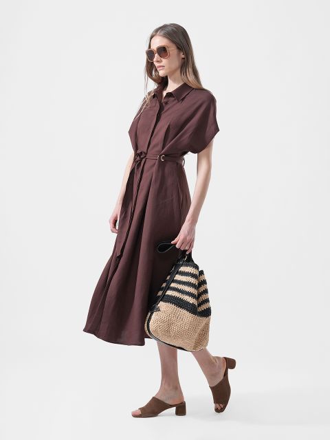 Frau trägt ein braunes Midikleid mit Gürtel, braune Gleitsandalen, eine Sonnenbrille und eine geflochtene Tragetasche aus Stroh mit schwarzen Streifen.