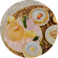 Buntes Geschirr mit Obstmotiven, rosa und lila Gläser und gelbe Früchte auf einem runden Holztisch mit einem rosa Tischset.