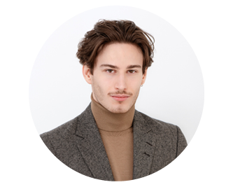 Junger Mann mit kurzen braunen Haaren, der einen grauen Blazer und einen hellbraunen Rollkragenpullover trägt und vor einem schlichten hellen Hintergrund posiert.