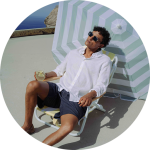 Ein Mann entspannt sich auf einem Liegestuhl unter einem gestreiften Sonnenschirm, in der Hand ein Glas Weißwein, in der Nähe eines Pools mit Blick auf das Meer im Hintergrund.