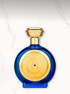 Ein runder blauer Parfümflakon mit goldenem Verschluss und einem kunstvollen goldenen Muster auf der Vorderseite, das sich vor einem weißen Marmorhintergrund abhebt.