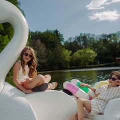 Zwei Kinder mit Sonnenbrillen entspannen sich auf einem schwanenförmigen Paddelboot auf einem See, umgeben von Bäumen unter einem sonnigen Himmel.
