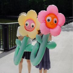Zwei Personen halten große aufblasbare Blumen mit lächelnden Gesichtern, die ihre eigenen Gesichter verdecken, während sie auf einer Brücke am Wasser stehen.