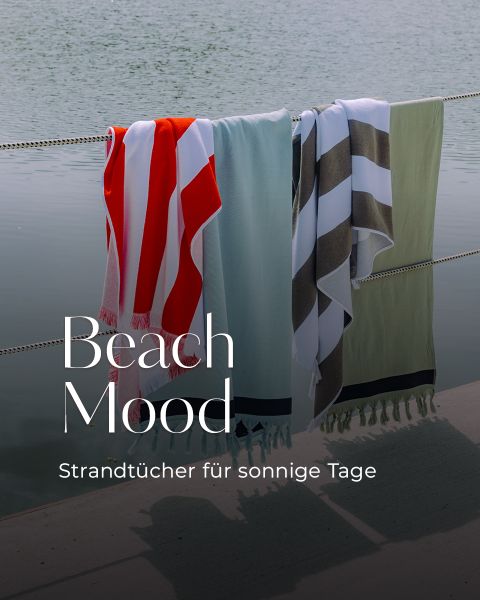 Drei bunte Strandtücher hängen an einer Leine am Wasser, über dem Bild ist der Text „Beach Mood. Strandtücher für sonnige Tage“ eingeblendet.