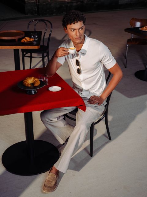 Mann in weißem Hemd und beiger Hose sitzt an einem Cafétisch mit rotem Tischtuch und trinkt Espresso, während auf dem Tisch Croissants liegen.