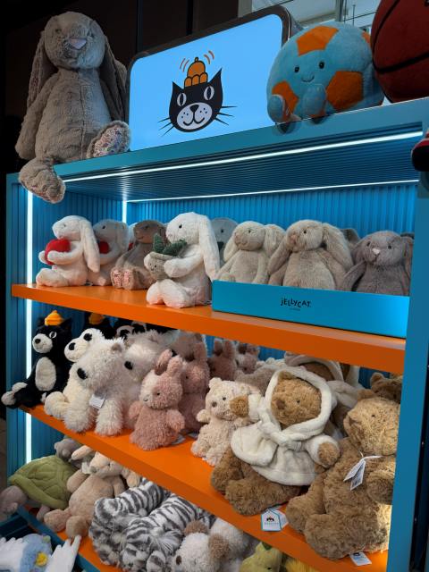 Display of colorful plush stuffed animals on blue and orange shelves in a toy store, including bunnies, bears, and a Jellycat sign/logo above.