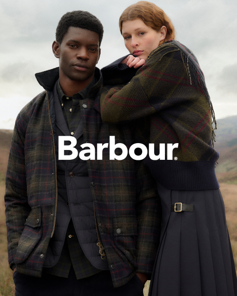Ein Mann und eine Frau in karierten Barbour-Jacken posieren im Freien, dicht beieinander vor einem bewölkten Himmel und einer hügeligen Landschaft. Das Barbour-Logo ist mittig platziert.