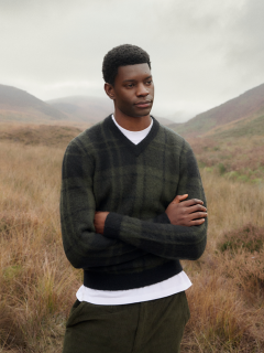 Ein Mann in einem karierten Pullover steht mit verschränkten Armen auf einer Wiese, im Hintergrund sind Berge und ein bewölkter Himmel zu sehen.