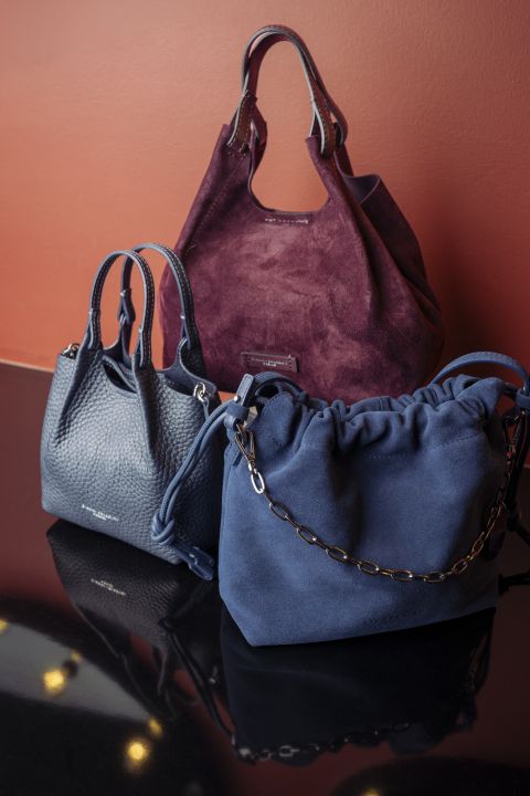 Three handbags displayed together: a burgundy suede tote, a blue textured leather tote, and a navy blue drawstring bag against an orange wall.