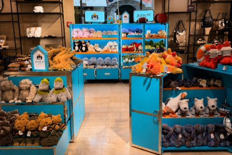 Ein hell beleuchtetes Geschäft mit blauen Regalen, in denen verschiedene Plüschtiere ausgestellt sind, darunter Hunde, Dinosaurier und Bären. Im Hintergrund hängen Handtaschen an Wandregalen.