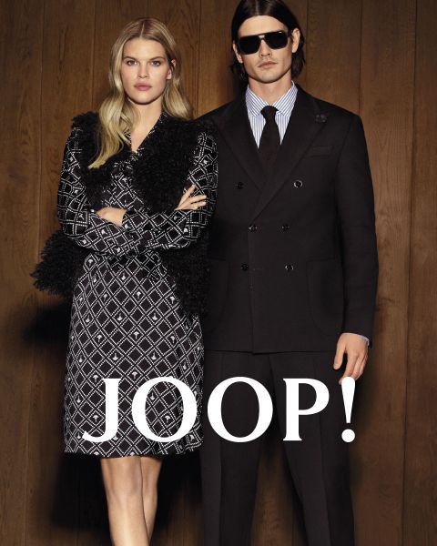 Eine Frau in einem gemusterten Kleid und ein Mann in einem schwarzen Anzug stehen nebeneinander vor einer Holzwand, auf der in fetten weißen Buchstaben „JOOP!“ steht.