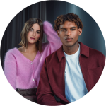 Zwei Personen posieren in einem Raum, eine trägt einen rosa Pullover, die andere eine weinrote Jacke; beide blicken direkt in die Kamera.