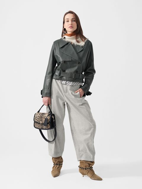 Eine Frau in einer schwarzen Lederjacke, einer karierten Hose und hellbraunen Stiefeln, die eine Handtasche mit Leopardenmuster in der Hand hält, steht vor einem schlichten weißen Hintergrund.