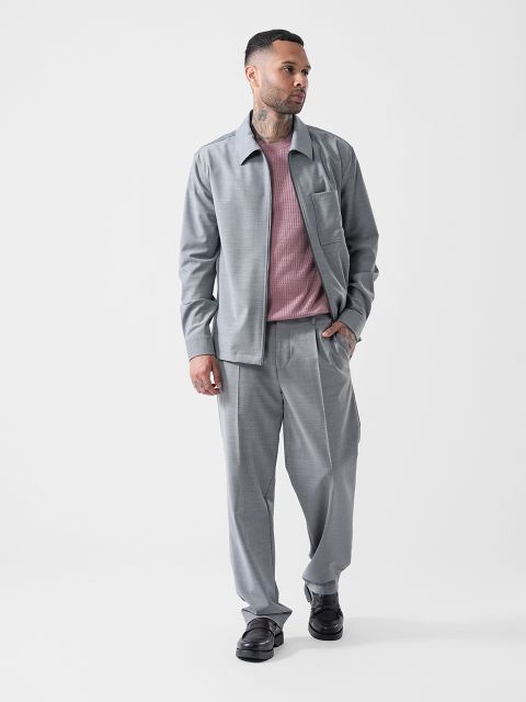 Ein Mann in einem grauen Anzug mit einem rosa Hemd, schwarzen Schuhen und der Hand in der Hosentasche, der vor einem schlichten weißen Hintergrund posiert.