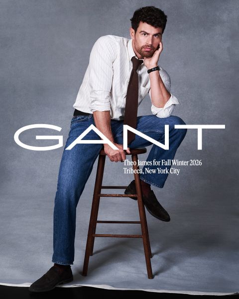 Ein Mann in gestreiftem Hemd, Krawatte und Jeans posiert auf einem Holzhocker vor einem grauen Hintergrund. Text: „GANT, Theo James für Herbst/Winter 2026, Tribeca, New York City.“.