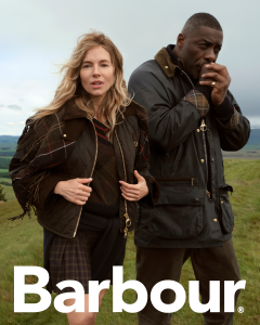 Eine Frau und ein Mann in Outdoor-Jacken stehen auf einem grasbewachsenen Hügel unter einem bewölkten Himmel. Unten steht in großen weißen Buchstaben das Wort „Barbour“.